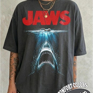 Op de afbeelding: Een antracietgrijs T-shirt met korte mouwen voorzien van het rode JAWS-filmlogo boven een afbeelding van een zwemmer aan het wateroppervlak en een grote haai met open bek die uit het diepblauwe water omhoog komt. Het shirt heeft een wijde pasvorm.
