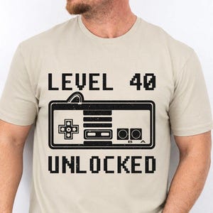 May include: A beige short-sleeve t-shirt featuring a black graphic print of a retro video game controller. The text above the controller reads LEVEL 40 and the text below reads UNLOCKED, both in a pixelated font style on the front of the shirt.