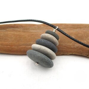May include: A handcrafted necklace featuring a pendant made of stacked, smooth river stones in shades of gray and beige. The stones are strung on a black cord, creating a natural and minimalist design. The pendant is approximately 2 inches long.