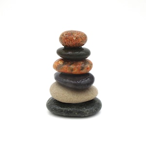 May include: A stack of six smooth, colorful stones arranged vertically. The stones vary in color, including shades of brown, black, gray, and tan. The arrangement suggests balance and harmony, set against a plain white background.