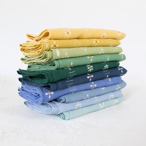 May include: A stack of folded square napkins in a gradient of colours ranging from yellow to light blue. Each napkin features a repeating pattern of small white daisy-like flowers with orange centres, and each has a frayed, raw edge finish.
