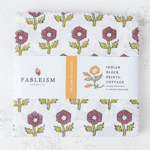 May include: A stack of fabric squares featuring a white background with a repeating pattern of maroon flowers and green leaves. A paper band wraps around the center with the text Fableism Supply Co, Indian Block Prints: Cottage, and 5 inch charm squares.