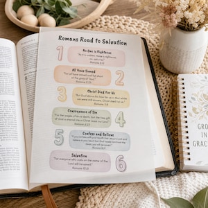 May include: A white paper sheet titled Romans Road to Salvation featuring six numbered steps in pastel colors. Each step includes a heading and a Bible verse. The sheet is placed on an open Bible, with a spiral notebook and a vase of dried flowers nearby.