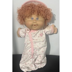 Pode incluir: Uma boneca Cabbage Patch Kids com cabelo de lã encaracolado castanho-claro, olhos azuis e rosto redondo. A boneca veste um pijama branco de manga comprida com padrão de corações cor-de-rosa, acabamento rosa, um laço pequeno e um logótipo CPK.