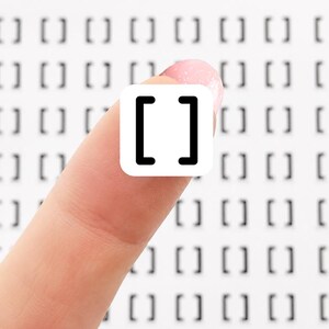 May include: A small, white square sticker with a black bracket symbol is held between fingers. The sticker is centered and in focus, with a grid of bracket symbols in the background. The background is white.