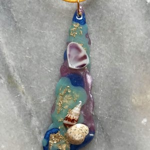 Puede incluir: Un collar con colgante artesanal con un diseño de resina en tonos azules, morados y turquesas. El colgante está adornado con conchas marinas y detalles de pan de oro, suspendido de un cordón amarillo.