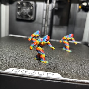 May include: Three small, multi-colored 3D printed figurines of a bipedal creature with a hunched posture, featuring a vibrant rainbow gradient of red, orange, yellow, green, and blue, standing on the textured black print bed of a Creality machine.