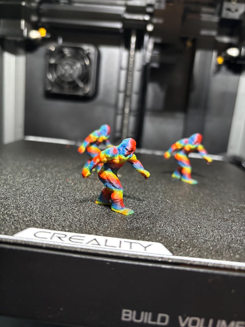 May include: Three small, multi-colored 3D printed figurines of a bipedal creature with a hunched posture, featuring a vibrant rainbow gradient of red, orange, yellow, green, and blue, standing on the textured black print bed of a Creality machine.
