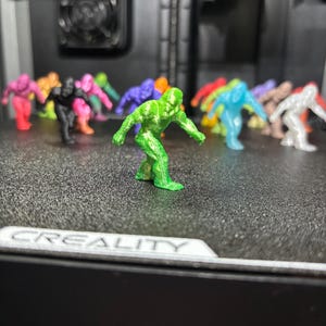 May include: A collection of small, 3D-printed humanoid figurines in various colors including green, black, pink, blue, orange, and white. The figures are posed in a forward-leaning stance and are arranged on a textured black build plate.