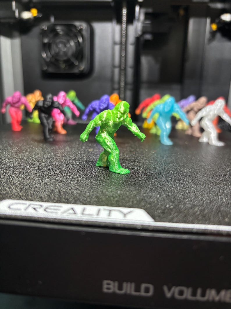 May include: A collection of small, 3D-printed humanoid figurines in various colors including green, black, pink, blue, orange, and white. The figures are posed in a forward-leaning stance and are arranged on a textured black build plate.