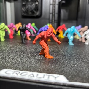 May include: A collection of small, 3D-printed figurines shaped like Bigfoot, rendered in various solid colors including red, orange, yellow, green, blue, purple, black, and white. The figures are arranged on a textured black surface with the text CREALITY.