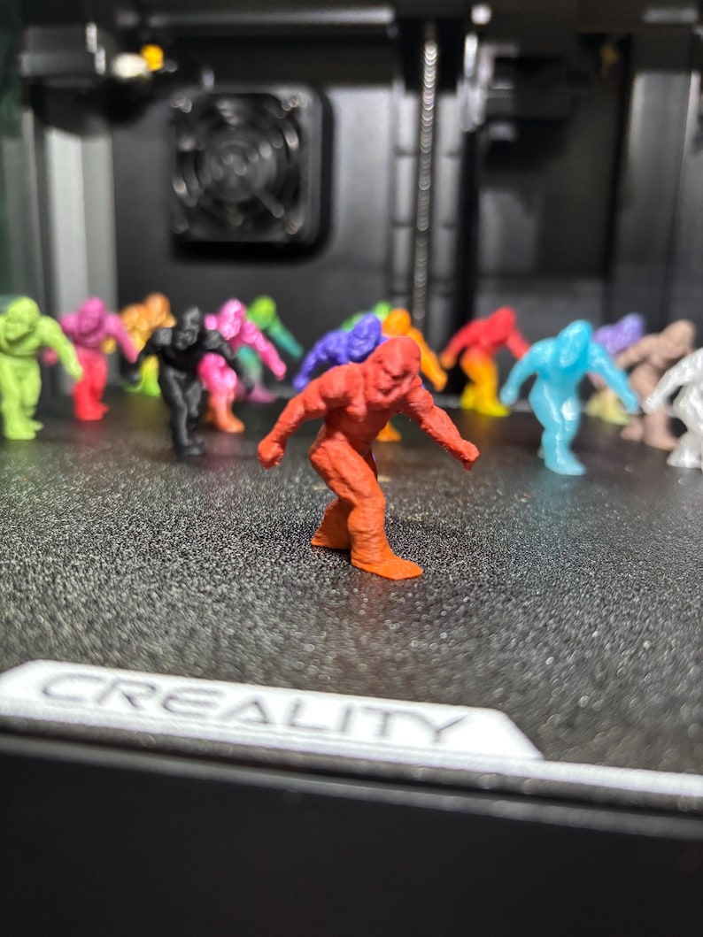 May include: A collection of small, 3D-printed figurines shaped like Bigfoot, rendered in various solid colors including red, orange, yellow, green, blue, purple, black, and white. The figures are arranged on a textured black surface with the text CREALITY.