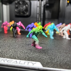 May include: A collection of small, 3D-printed humanoid figurines in various solid and gradient colors, including a prominent foreground figure featuring a green-to-pink color transition, all standing in a crouched, forward-leaning pose.
