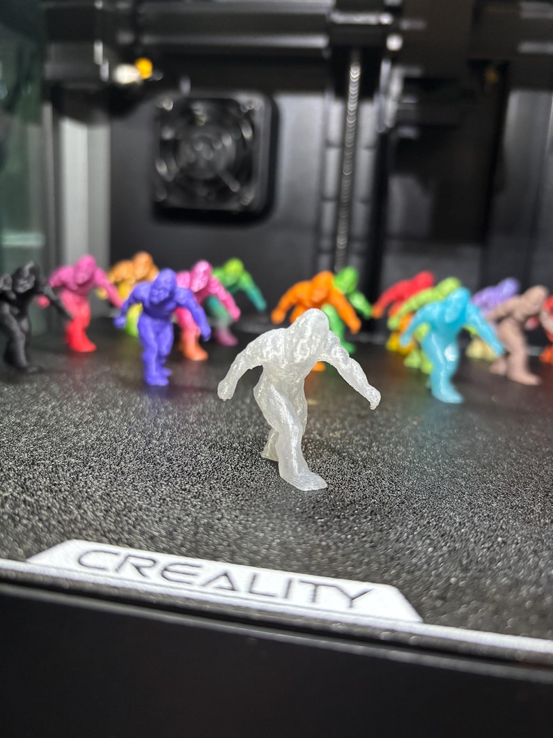 May include: A collection of small, 3D-printed humanoid figurines in various colors, including translucent, purple, pink, orange, green, blue, and black. The figures are in a dynamic, crouching pose. The word CREALITY is printed on the surface.
