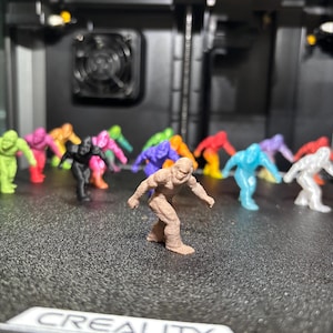 May include: A collection of small, 3D-printed figurines shaped like Bigfoot, rendered in a variety of solid colors including brown, lime green, pink, orange, black, purple, blue, and white, arranged in a group on a textured black surface.