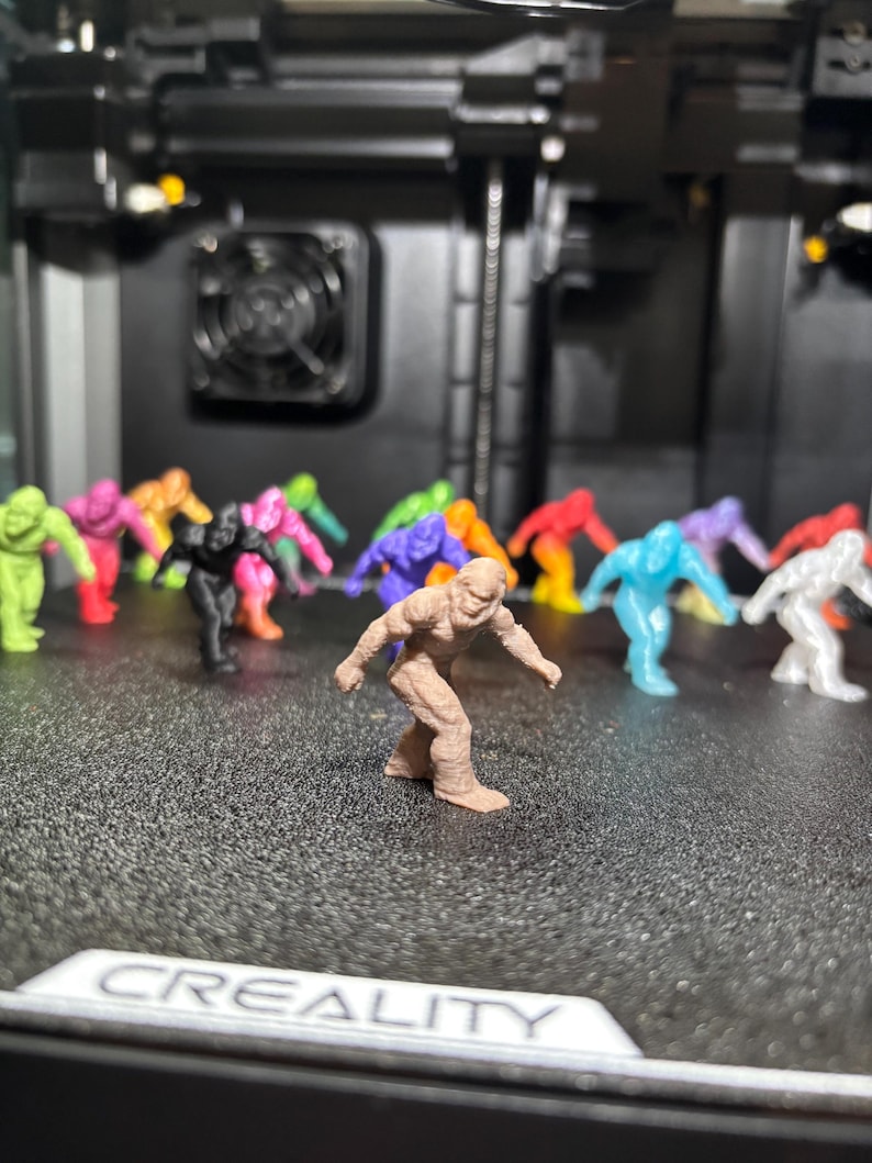 May include: A collection of small, 3D-printed figurines shaped like Bigfoot, rendered in a variety of solid colors including brown, lime green, pink, orange, black, purple, blue, and white, arranged in a group on a textured black surface.
