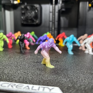 May include: A collection of small, 3D-printed humanoid figurines in various solid and gradient colors, including purple, yellow, blue, black, and pink. Each figure is in a dynamic, crouching pose with arms extended. The word CREALITY is visible.