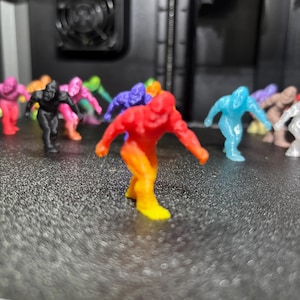 May include: A collection of small, 3D-printed gorilla figurines in various colors, including a prominent red and yellow gradient figure in the foreground, with others in black, blue, pink, and tan scattered across a textured black surface.