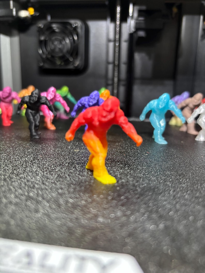May include: A collection of small, 3D-printed gorilla figurines in various colors, including a prominent red and yellow gradient figure in the foreground, with others in black, blue, pink, and tan scattered across a textured black surface.
