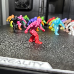 May include: A collection of small, 3D-printed plastic figurines shaped like Bigfoot or Sasquatch, displayed in various colors including red, pink, black, blue, green, orange, and white, arranged on a textured black surface with the text CREALITY.