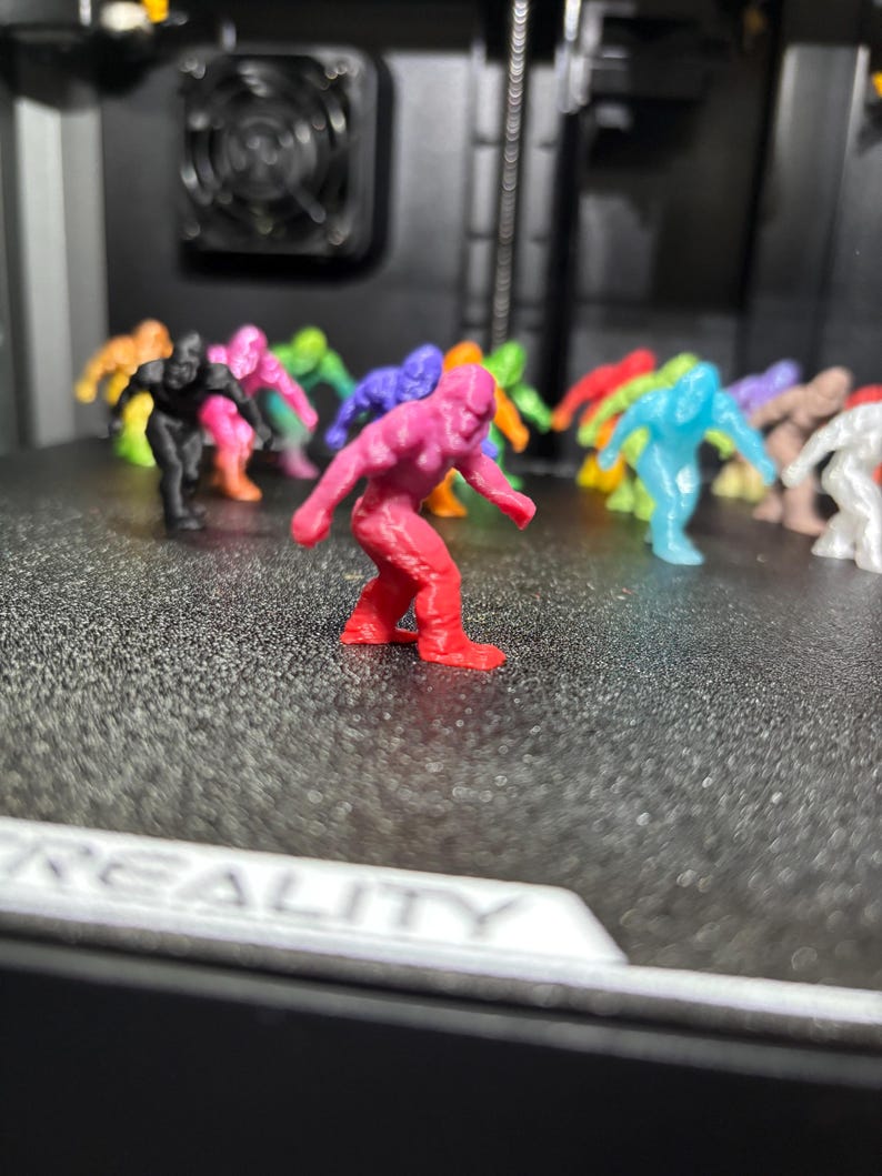 May include: A collection of small, 3D-printed plastic figurines shaped like Bigfoot or Sasquatch, displayed in various colors including red, pink, black, blue, green, orange, and white, arranged on a textured black surface with the text CREALITY.