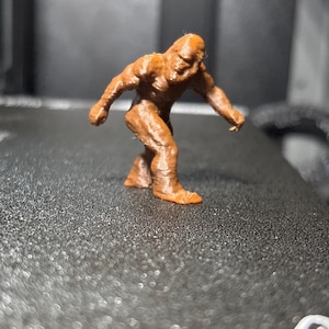 May include: A small, brown, three-dimensional printed figurine of a bipedal creature with a hunched posture, muscular build, and textured surface, depicted in a walking stance with one arm extended forward and the other arm positioned behind.
