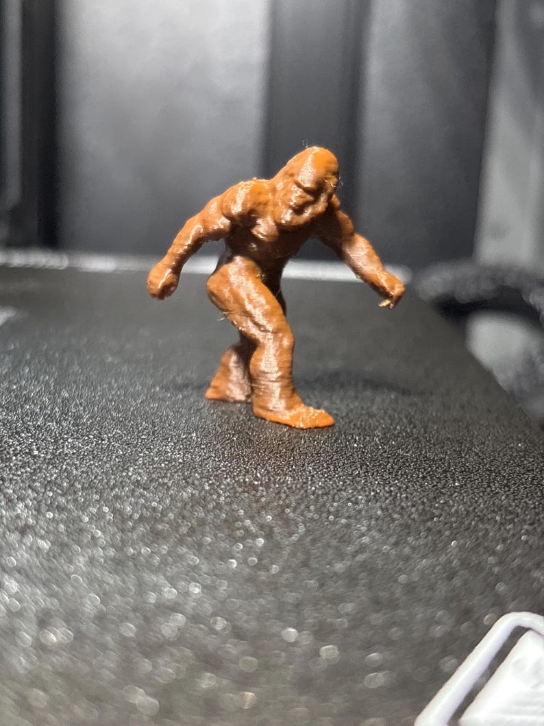 May include: A small, brown, three-dimensional printed figurine of a bipedal creature with a hunched posture, muscular build, and textured surface, depicted in a walking stance with one arm extended forward and the other arm positioned behind.