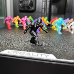 May include: A collection of small, 3D-printed figurines shaped like Bigfoot, featuring a black figure in the foreground and various colorful translucent and opaque figures in the background, all resting on a textured black printer build plate.