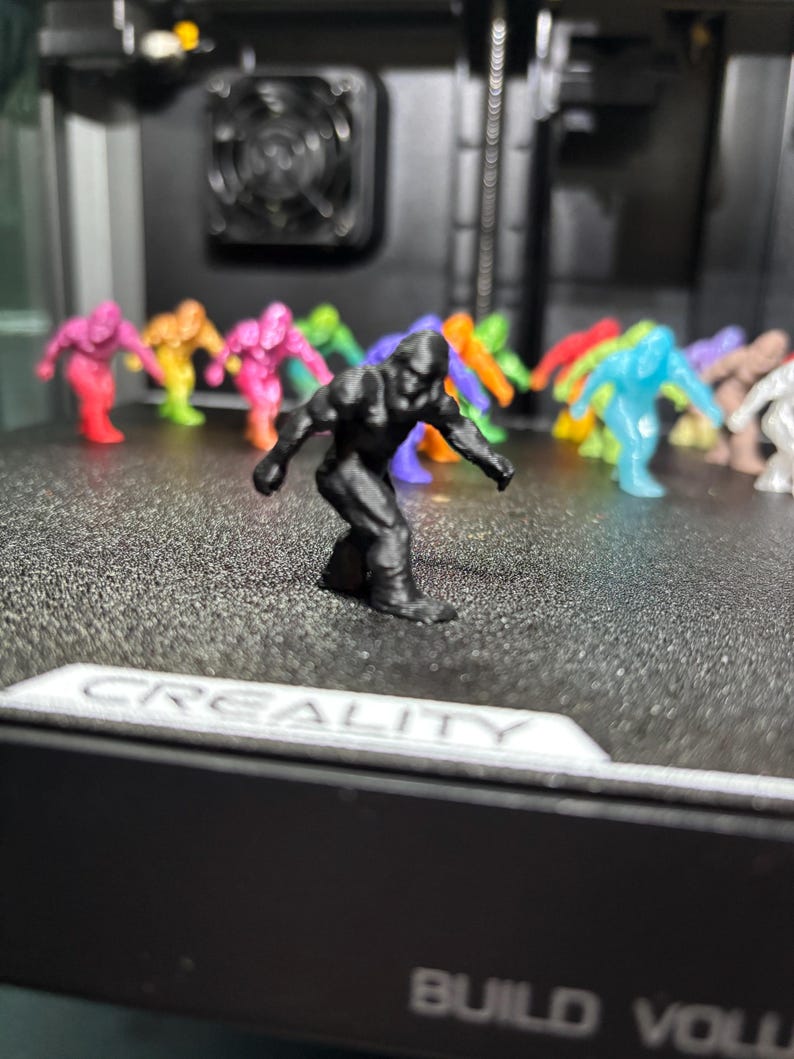 May include: A collection of small, 3D-printed figurines shaped like Bigfoot, featuring a black figure in the foreground and various colorful translucent and opaque figures in the background, all resting on a textured black printer build plate.