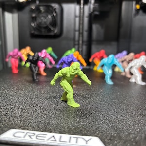 May include: A collection of small, 3D-printed humanoid monster figurines in various solid colors including lime green, black, pink, orange, purple, blue, and white, arranged in a group with one lime green figure standing in the foreground.