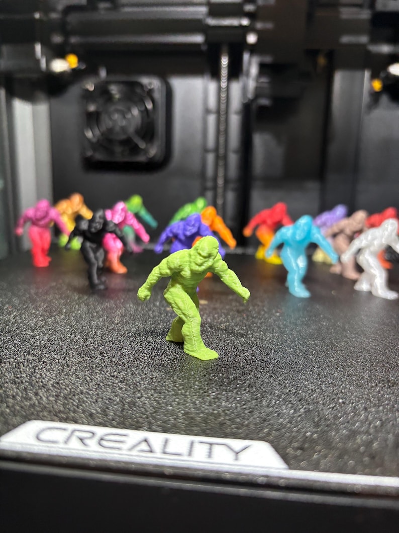 May include: A collection of small, 3D-printed humanoid monster figurines in various solid colors including lime green, black, pink, orange, purple, blue, and white, arranged in a group with one lime green figure standing in the foreground.