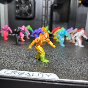 May include: A collection of small, 3D-printed humanoid figurines in various solid and gradient colors, including orange, yellow, green, pink, black, blue, purple, and white, standing in a group on a textured black surface with the text CREALITY.