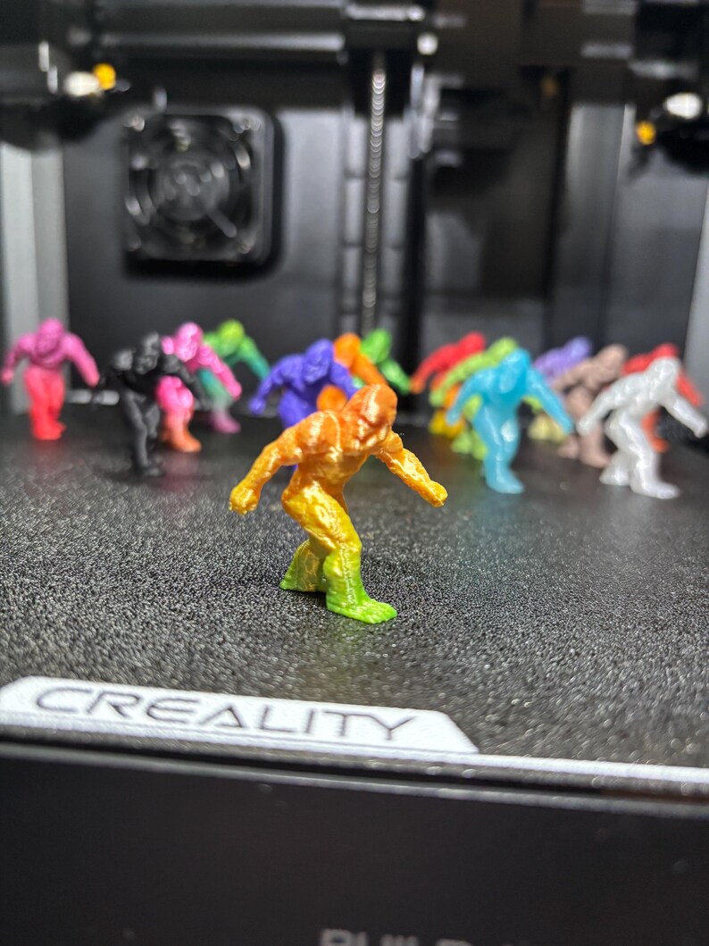 May include: A collection of small, 3D-printed humanoid figurines in various solid and gradient colors, including orange, yellow, green, pink, black, blue, purple, and white, standing in a group on a textured black surface with the text CREALITY.