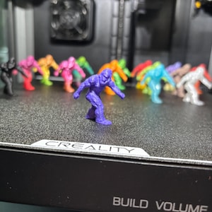 May include: A collection of small, 3D-printed figurines shaped like gorillas in various solid colors, including purple, black, pink, orange, blue, green, and white, arranged on a black textured surface with the text CREALITY BUILD VOLUME.