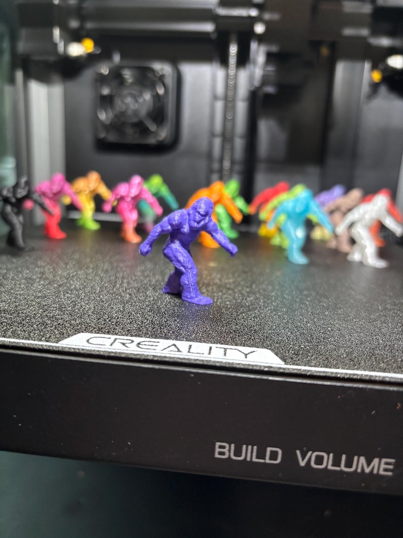 May include: A collection of small, 3D-printed figurines shaped like gorillas in various solid colors, including purple, black, pink, orange, blue, green, and white, arranged on a black textured surface with the text CREALITY BUILD VOLUME.
