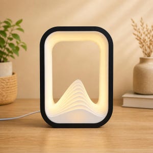 May include: A modern, rectangular LED lamp with a black frame and a white interior. The lamp features a mountain-shaped design in the center, with layered, illuminated pages. The lamp is plugged into a wall outlet and sits on a wooden surface.
