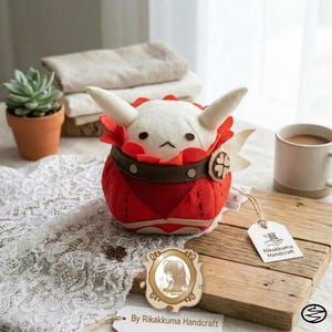 May include: A plush toy featuring a white creature with two horns, nestled inside a red, rounded, fabric-based container with a brown belt-like strap. The toy has small black eyes and a simple mouth. A white tag reads Rikakkuma Handcraft.