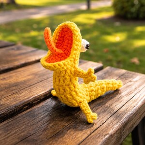 May include: A handmade yellow crochet duckling toy with orange accents inside the hood and beak. The duckling is sitting on a wooden bench, with its arms and legs outstretched. The background is blurred, showing green grass and trees.