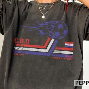 May include: Dark gray t-shirt featuring a soccer ball with blue and white streaks, the letters "C.R.O", and the Croatian flag. The shirt also includes the text "EST. 1912" and "WORLD CUP 2026".