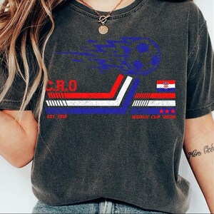 May include: Dark gray t-shirt featuring a soccer ball graphic with blue flames. The shirt includes the text "CRO" and "WORLD CUP 2026" with the Croatian flag design. The shirt also has the text "EST. 1912".
