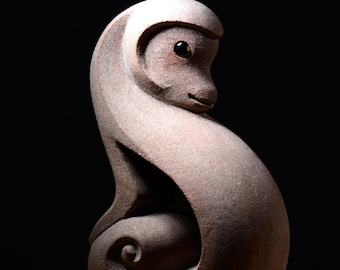 May include: A smooth, light brown stone sculpture of a stylized monkey with a curled tail. The figure features a rounded head, a prominent brow ridge, and a single dark, polished eye. The body curves into a spiral shape at the base.