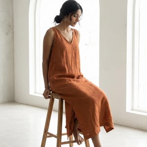以下が含まれることがあります： 無地のバーントオレンジ色のノースリーブミディ丈ドレス。Vネックライン、リラックスフィット、裾のサイドスリットが特徴です。生地にはリネン特有の目に見える質感のある織り目があり、ふくらはぎの中間までの長さです。