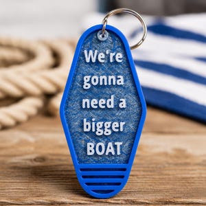 May include: Blue keychain with the text "We're gonna need a bigger BOAT" in white. The keychain has a retro motel style design and a silver key ring. The background includes a rope and a blue and white striped fabric.