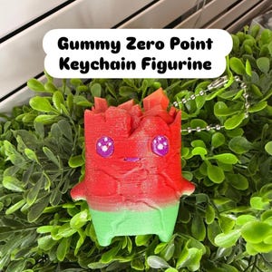 May include: A Gummy Zero Point Keychain Figurine featuring a red top and green bottom with a textured surface. The character has two purple eyes with white highlights and a small mouth. A silver metal ball chain is attached to the top of the figure.