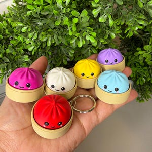 May include: A collection of six colourful dumpling keychains, each featuring a smiling face with pink cheeks. The dumplings are red, white, yellow, light blue, purple, and magenta, and each sits inside a small, tan, circular steamer basket base.