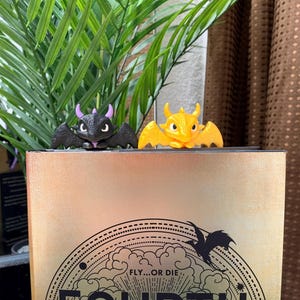 May include: Two small dragon-shaped bookmarks perched on top of a gold book cover. One dragon is black with purple horns, and the other is bright yellow. The book cover features a circular graphic with the text FLY...OR DIE.