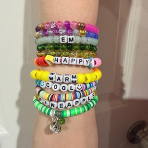 May include: A stack of colorful beaded bracelets. The bracelets feature various colors, including pink, green, yellow, and blue. Some bracelets have letter beads spelling out words like "EM," "HAPPY," "WARM," "COOL," and "PINEAPPLE." A silver charm hangs from one bracelet.
