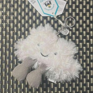 May include: A white, fluffy cloud-shaped keychain toy with grey corduroy feet and a smiling face. The keychain has a silver ring and a tag that reads "Jellycat". The background is a woven pattern.