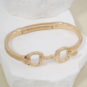 May include: A gold-toned metal hinged bracelet featuring a textured, coiled spring section on one side and a central horse-bit style link closure. The bracelet has a smooth, polished finish and a curved, rigid band structure.