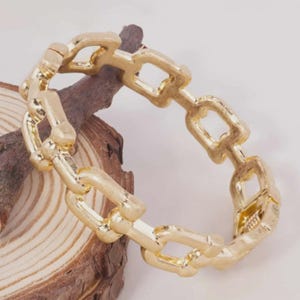 May include: A gold-toned metal bracelet featuring a series of interconnected rectangular links. The bracelet has a polished finish and a visible hinged clasp mechanism on one side, creating a continuous chain-link design for the wrist.
